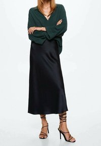 Femme portant un blouse vert foncé à manches amples, une jupe midi en satin noir, et des sandales à talons hauts noires à lacets, se tenant les bras croisés.