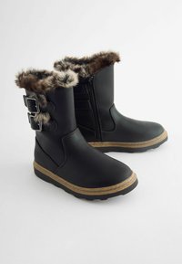 Bottes noires imperméables avec bordure en fausse fourrure et boucles. Présentent une fermeture éclair sur le côté et une semelle en caoutchouc texturée avec une semelle intermédiaire beige.