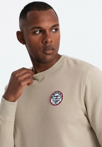 Sudadera beige de cuello redondo con un acabado texturizado que presenta un parche redondo en el lado izquierdo del pecho con detalles en negro y rojo.