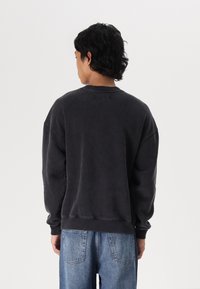 Črna oversized dukserica z ovratnikom na krog, spuščenimi rameni in rebrastimi manšetami, v kombinaciji s svetlo modrimi jeans hlačami. Gladka tekstura.