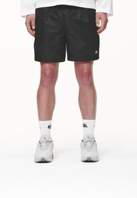 Persona che indossa pantaloni corti neri, calze bianche a girocollo con loghi e sneakers bianche, in piedi con le mani rilassate ai lati su uno sfondo bianco.