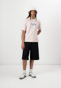 Roza bombažna majica s modrim napisom, skupaj s črnimi kratkimi hlačami. Oblečena z bež kapico in sivimi belimi teniskami z črnimi črtami.