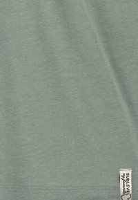 Light green T-shirt made of soft fabric, featuring a simple design and a visible label with "SUBTLE" text in the corner.