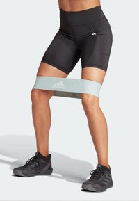 Weerstandsband in lichtblauw, om de dijen geplaatst, met Adidas-logo. Zwarte high-waisted shorts en zwarte sportschoenen worden gedragen.