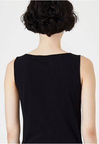 Person med kort, lockigt mörkt hår iförd en ärmlös svart topp, sedd bakifrån mot en enkel vit bakgrund.