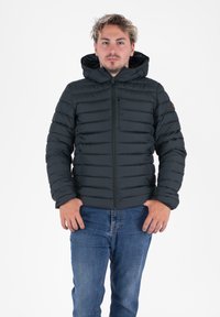 Man in een zwarte gewatteerde hoodie en blauwe jeans, staand met het gezicht naar voren en een neutrale uitdrukking.