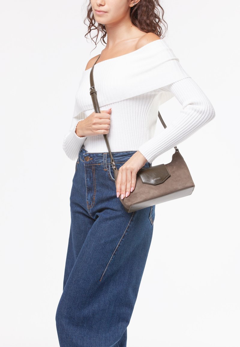 Mała brązowa torebka na przekór z zamszowego materiału, z klapą i długim regulowanym paskiem, noszona przez osobę w białym swetrze.