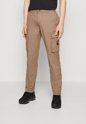 Person med beige cargo-byxor med fickor och svarta sneakers, som står mot en enfärgad ljus bakgrund.