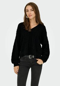 Pull en maille noir avec un col en V, motif texturé et manches longues, associé à un jean noir et une ceinture. Style décontracté et coupe relax.