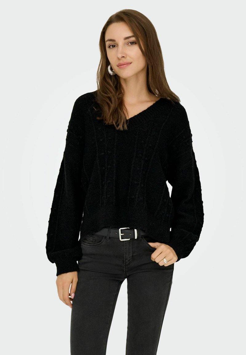 Pull en maille noir avec un col en V, motif texturé et manches longues, associé à un jean noir et une ceinture. Style décontracté et coupe relax.