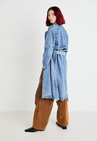 Trench coat in denim azzurro con maniche lunghe e vita regolabile, abbinato a pantaloni a gamba larga marroni e stivali neri con punte appuntite.