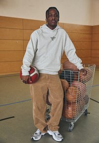 Felpa grigia con logo, pantaloni di corduroy color cammello e sneakers argento. Tiene una palla da basket rossa e bianca accanto a un carrello di metallo pieno di palloni.