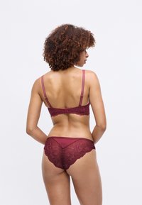 Donna con capelli ricci che indossa un reggiseno e una mutandina in pizzo bordeaux coordinati, di spalle contro uno sfondo bianco neutro.