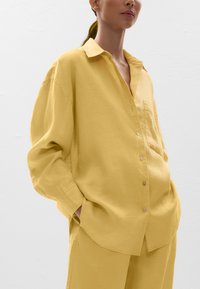 Chemise boutonnée en lin jaune avec une coupe décontractée, épaules basses et une poche poitrine. Texture lisse avec un design légèrement surdimensionné.