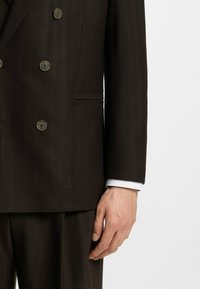 Giacca di completo doppiopetto marrone scuro con motivo a spina di pesce, caratterizzata da grandi bottoni, tasche laterali e un polsino di camicia bianca visibile nella manica.
