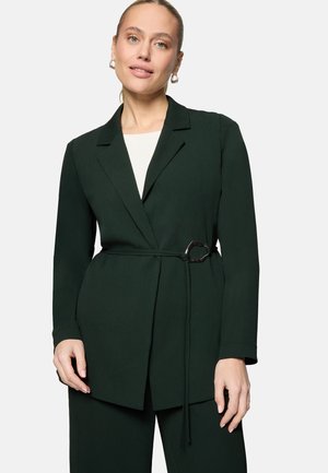 Vrouw die een donkergroene blazer met riem en bijpassende broek draagt, poserend tegen een eenvoudige lichte achtergrond.