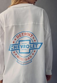 Cămașă albă cu mâneci lungi, având un logo vintage Chevrolet. Designul include text roșu și albastru, elemente circulare și accente cu stele.