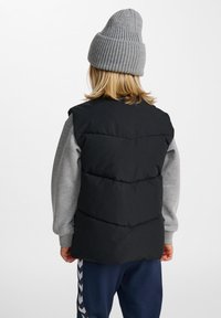 Schwarze, gepolsterte Weste mit einem gesteppten Design, getragen über einem grauen Langarmshirt. Die Person trägt eine gerippte graue Mütze und marineblaue Hose.