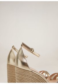 Sandales à talon en or métallique avec une plateforme en corde, bout pointu et bride de cheville réglable avec une boucle circulaire. Texture lisse.