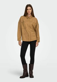 Tan faux suede oversized button-up shirt met twee borstzakken, gecombineerd met zwarte skinny jeans en bruine kniehoge laarzen.