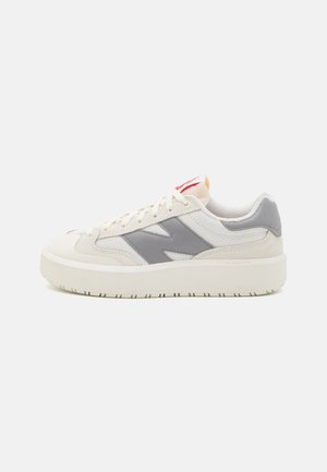 Zapatilla baja en blanco y gris con suela gruesa, superposiciones de malla y gamuza, y cordones blancos, vista de lado sobre un fondo liso.