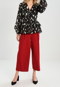 Blusa peplum nera con motivo a foglia dorata, scollatura a V profonda e maniche a palloncino, abbinata a pantaloni larghi rossi e tacchi alti neri.