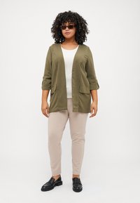 Cardigan verde oliva cu două buzunare frontale, mâneci trei sferturi, peste un top alb, asortat cu pantaloni bej și mocasi negri.