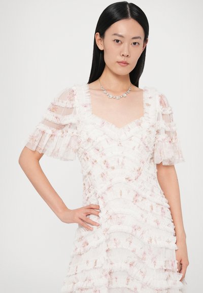 Biele šaty s volánikovými detailmi, priehľadnými krátkymi rukávmi a jemnými kvetinovými vzormi. Nosené s náhrdelníkom s drahokamami, ktorý má elegantný výstrih.