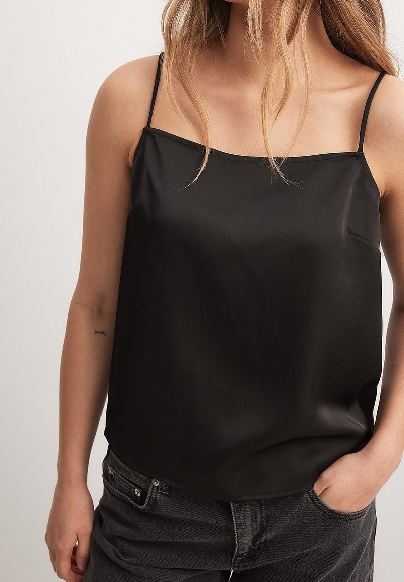 Camisola nera in satin con sottili spalline, caratterizzata da una vestibilità ampia e una superficie liscia, abbinata a jeans scuri nella parte inferiore.