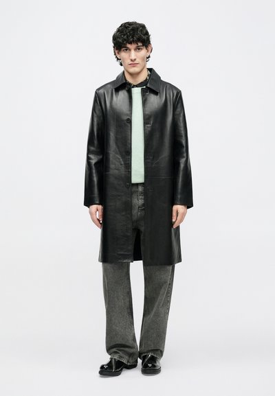 Noor mees seisab, kandes musta nahast pikka mantlit, mintrohelist kampsunit, musta mustrilist särki, pleekinud halli teksapükse ja musti kingi.