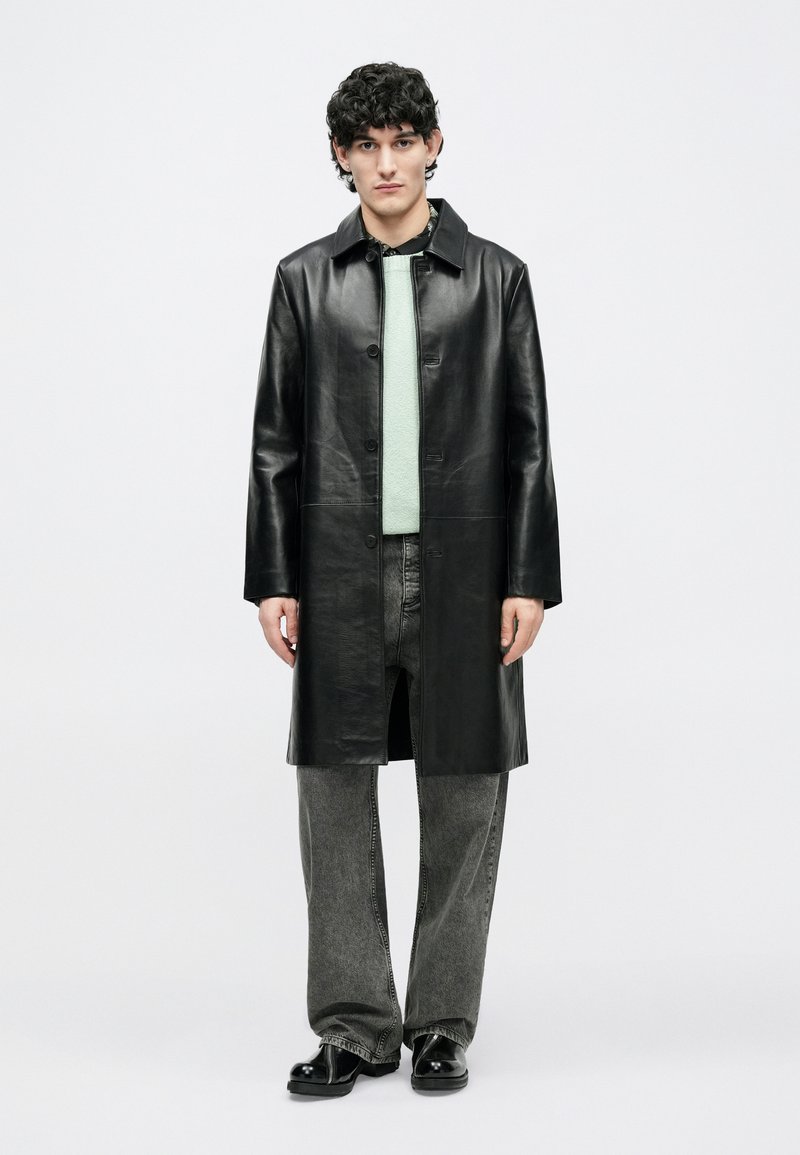 Jeune homme debout portant un long manteau en cuir noir, un pull vert menthe, une chemise noire à motifs, un jean gris délavé et des chaussures noires.