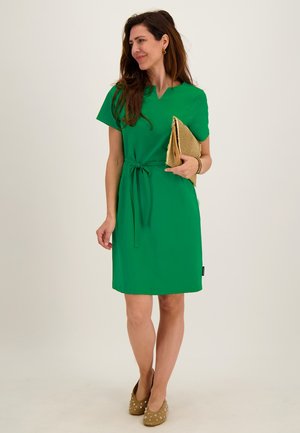 Vrouw in groene jurk tot op de knie, houdt een beige clutch vast, draagt bruine hakken met versieringen, staand tegen een effen achtergrond.