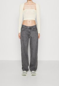 Kvinna som bär en krämfärgad stickad långärmad kort topp, lösa mörkgrå jeans och vita sneakers, stående mot en enkel bakgrund.