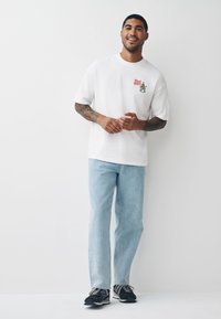 Balta oversize T-krekls ar krāsainu logotipu un grafiku, kombinēts ar gaiši ziliem džinsiem un melnām sporta kurpēm ar baltām detaļām.