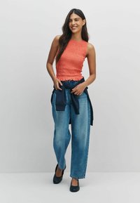 Korallikarva kudumitopp korseldatud tekstuuriga, kombineeritud helesiniste laiade lahjemate teksapükstega ja tumesinise vöökohale seotud kampsuniga. Mustad kingad.