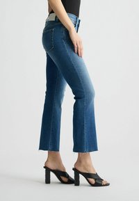 Blaue Denim-Jeans mit ausgefranstem Saum, mittelhohem Bund und leichter Ausbleichung. Kombiniert mit schwarzen Absatzsandalen mit gemusterten Blockabsätzen.