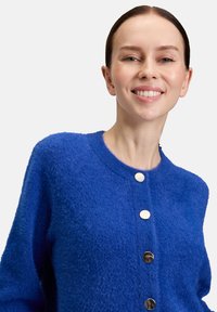 Jonge vrouw met donker haar glimlacht, draagt een felblauwe zachte cardigan met drie glanzende metalen knopen op een lichte achtergrond.