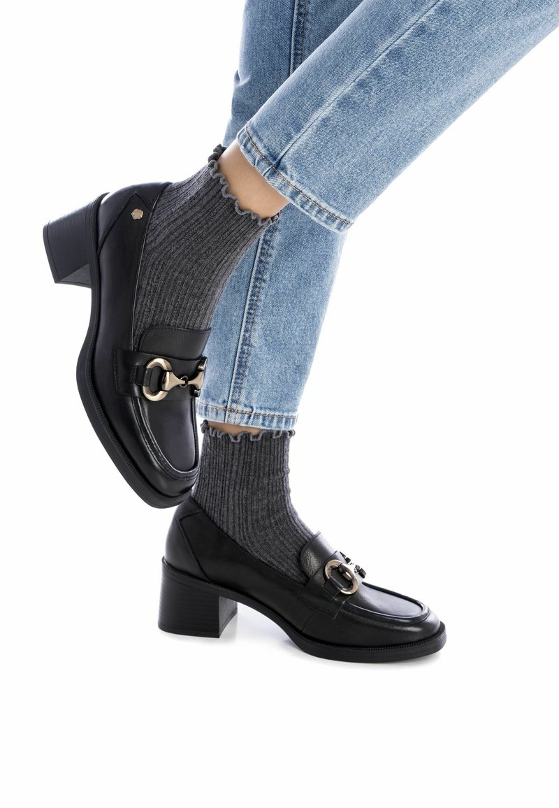Zapatos de cuero negro con tacón bloque y punta redondeada, con herrajes decorativos en dorado, usados con calcetines de rayas grises y combinados con jeans de mezclilla azul claro.