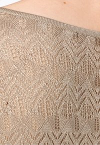 Tejido de punto beige con un delicado patrón de zigzag y una textura de tejido abierto. El escote está terminado con un suave borde acanalado.