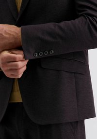 Mann, der den Ärmel eines dunklen, strukturierten Blazers mit vier schwarzen Knöpfen über einem senfgelben Strickpullover anpasst.