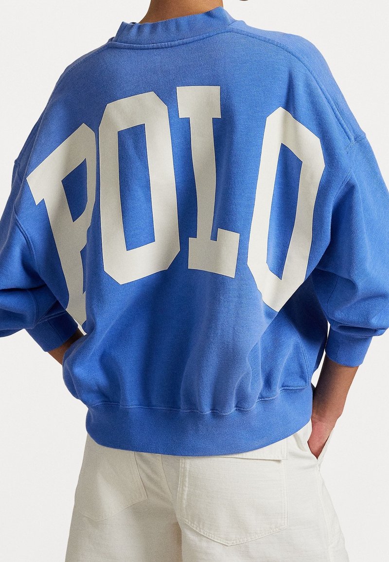 Personne portant un sweat-shirt bleu avec de grandes lettres blanches "POLO" dans le dos et un pantalon clair avec les mains dans les poches.