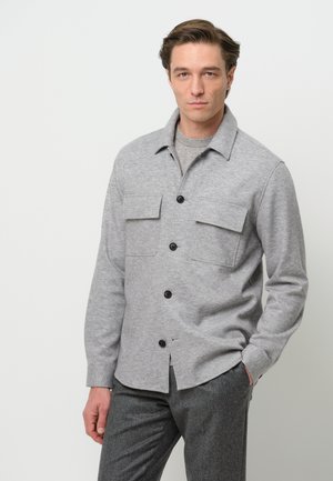 Grijze button-up jas van zachte stof met twee borstzakken, zwarte knopen en een klassieke kraag. G gedragen met grijze broek.