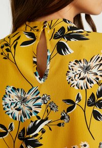 Woman wearing mustard yellow top with black, white, and blue floral pattern, featuring a buttoned keyhole closure at the back neckline.