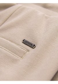Gros plan sur un tissu beige texturé avec une petite étiquette en métal marquée "ombre" cousue près d'une couture.