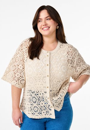 Beige blonder skjorte med geometriske mønstre, korte ærmer og knaplukning foran. Blød tekstur med bølget kant, båret over blå denim.