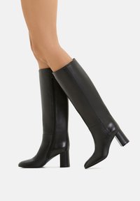 Stivali neri alti fino al ginocchio realizzati in pelle liscia, dotati di una cerniera laterale e di un tacco quadrato. Il design è elegante e aderente.