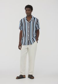 Camicia a maniche corte con bottoni, a strisce alternate nere e azzurro chiaro; abbinata a pantaloni bianchi larghi e sandali neri.