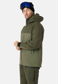 Giacca impermeabile verde oliva con cappuccio, dotata di tasche con zip e polsini regolabili. Abbinata a pantaloni utility coordinati.