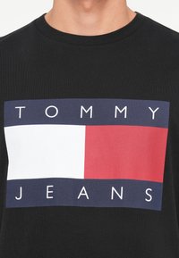 Musta t-särk, millel on ristkülikukujuline logo, kus ülal on "TOMMY" valge ploki kohal, all on punane plokk ja "JEANS" valge tekstiga.