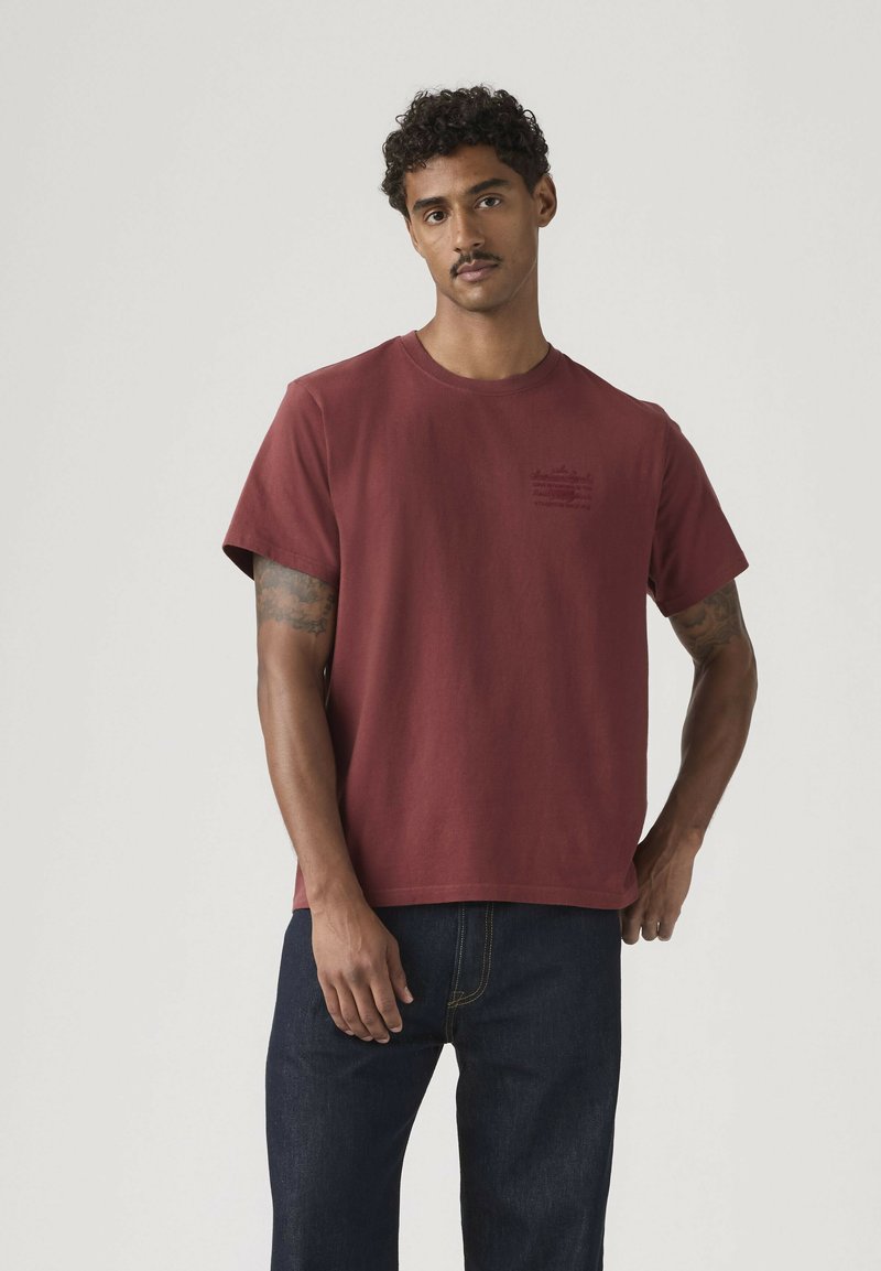 Mand med krøllet hår og overskæg iført en ensfarvet bordeaux kortærmet T-shirt og mørkeblå jeans, stående mod en lys baggrund.
